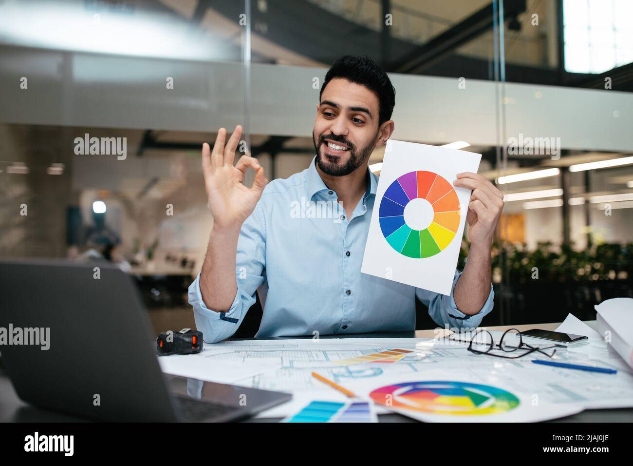 Fröhlicher junger islamischer Ingenieur-Designer mit Bart zeigt Farbkarte und OK-Schild mit Hand zum Laptop Stockfoto