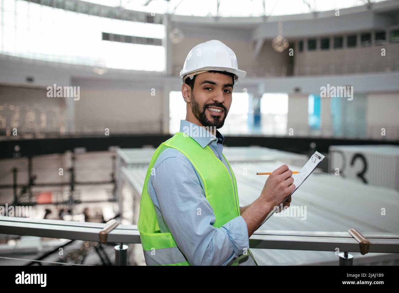 Frohes junger islamischer Profi-Ingenieur-Baumeister mit Bart in Schutzkleidung und Hut macht Notizen Stockfoto