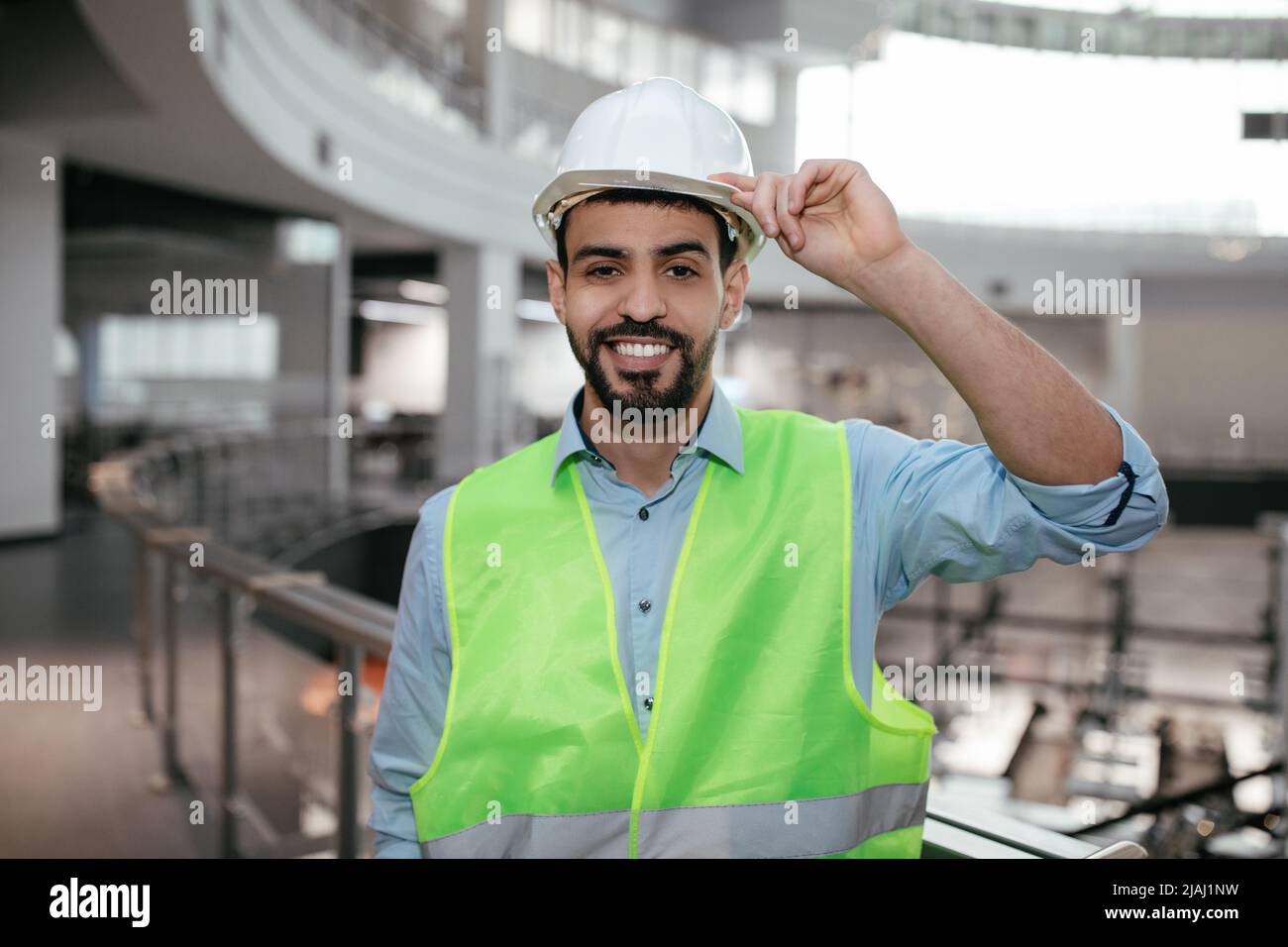 Happy Millennial Middle Eastern Bauingenieur mit Bart in Schutzkleidung berührt Hut mit der Hand Stockfoto