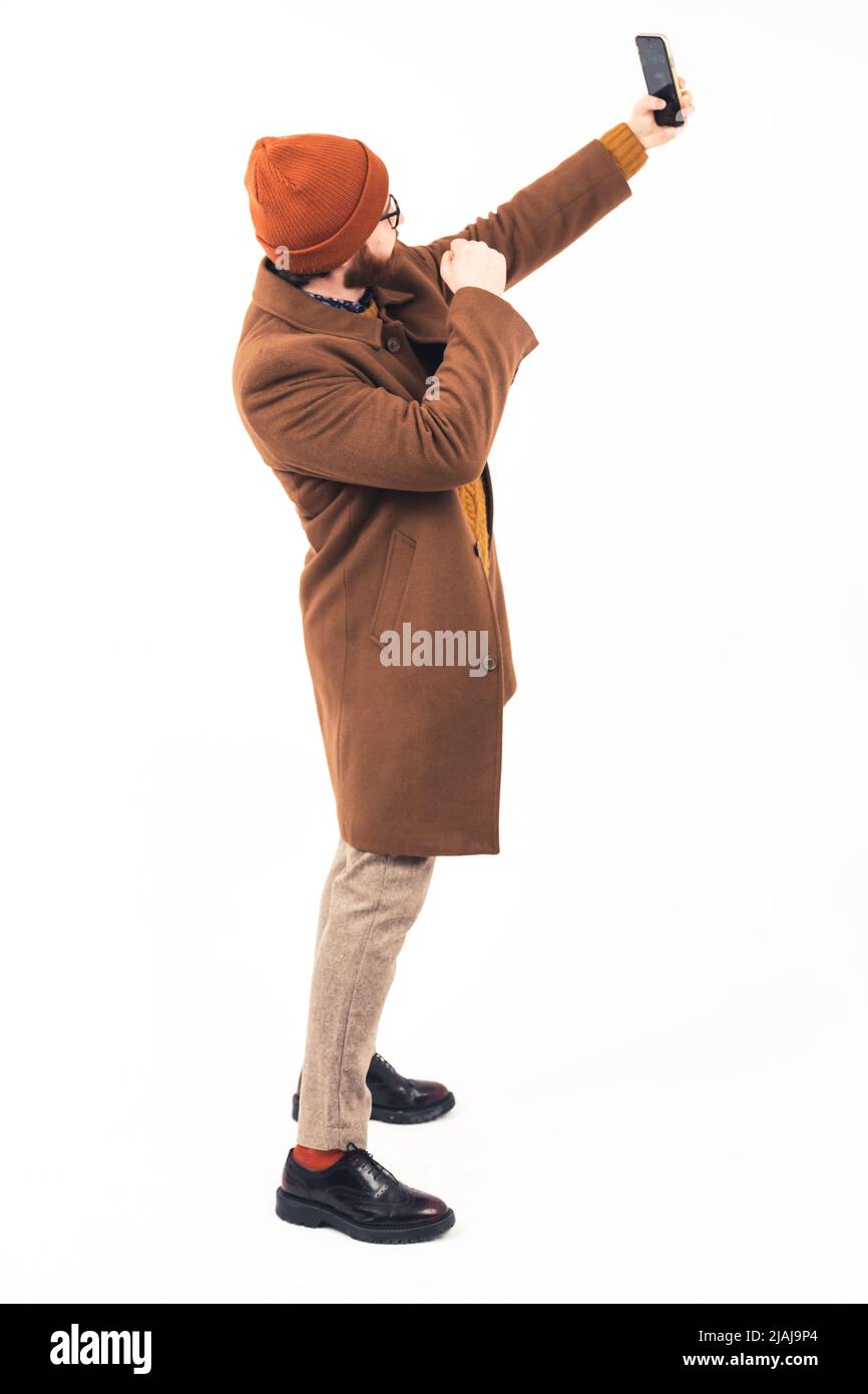 Käse. Vertikale Ganzkörperaufnahme eines nicht erkennbaren kaukasischen männlichen Hipsters, der für sein eigenes Selfie posiert, das mit einem modernen Smartphone aufgenommen wurde. Weißer Hintergrund. Hochwertige Fotos Stockfoto