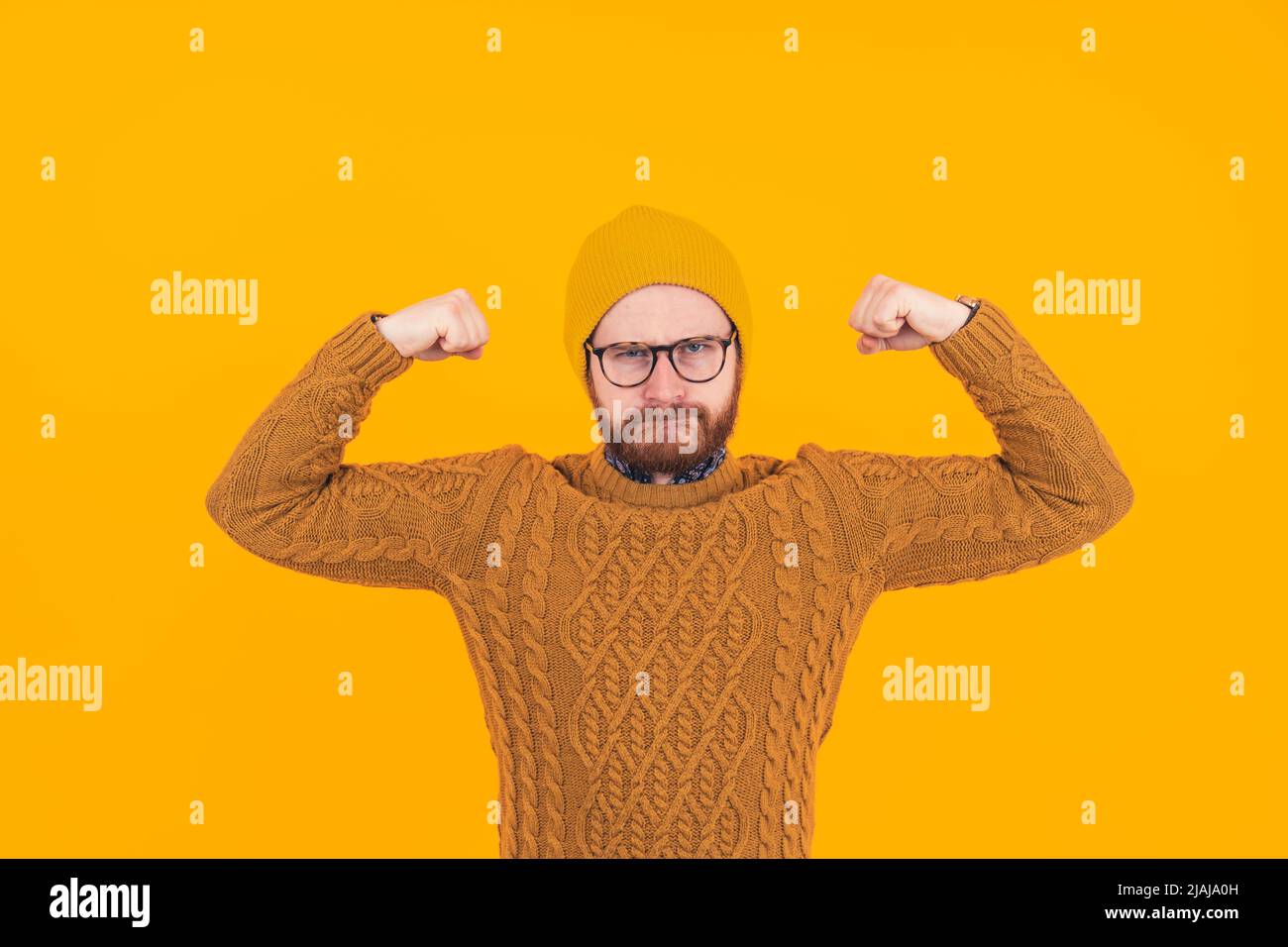 Lustiger europäischer bärtiger Hipster in einer Beanie spöttisch Bodybuilder durch Straffen seiner Bizeps, während das Tragen eines gestrickten Pullovers. Isoliertes Studio auf gelbem Hintergrund. Hochwertige Fotos Stockfoto