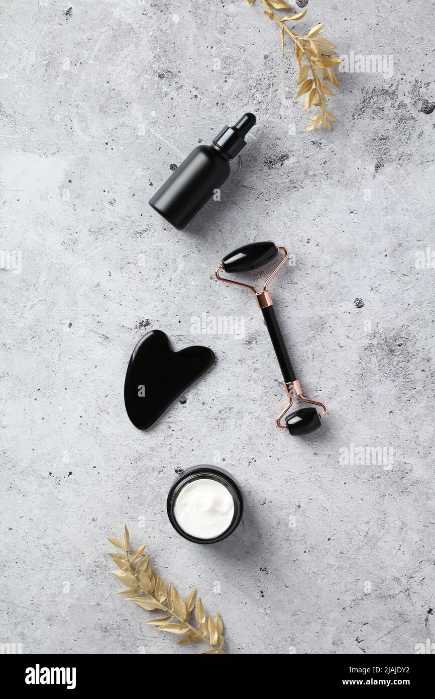 Obsidian Stone Face Roller, gua Sha, Serum Tropfflasche, Feuchtigkeitscreme Creme Glas und goldenen Zweigen auf Beton Hintergrund. Gesichtshautbehandlung, Anti Stockfoto