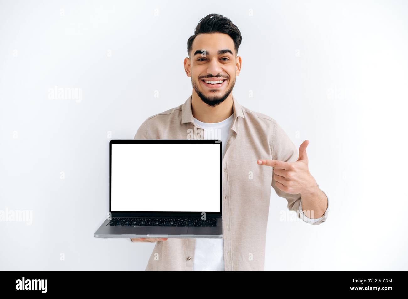 Fröhlicher arabischer oder indischer Mann in Gelegenheitskleidung, Freiberufler oder Student, hält einen offenen Laptop mit leerem Mock-up-Raum und zeigt den Finger darauf, steht auf weißem, isoliertem Hintergrund, schaut auf die Kamera, lächelt Stockfoto