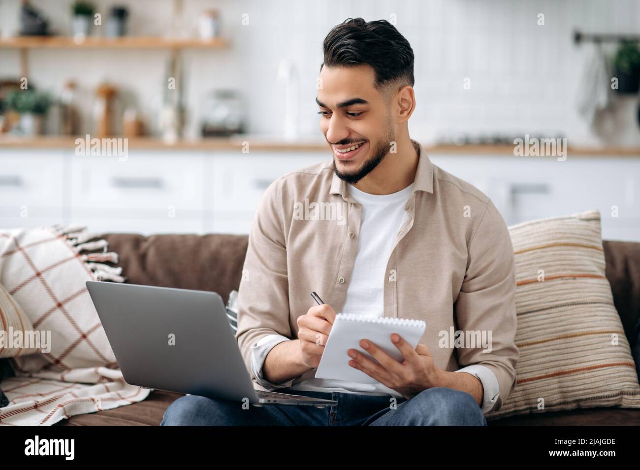 Ein positiver intelligenter inder oder araber, der sich Notizen macht, während er an einem Projekt arbeitet oder online lernt, verwendet einen Laptop, sitzt auf einem Sofa im Wohnzimmer, stöbert im Internet nach Informationen und bereitet einen Bericht vor Stockfoto