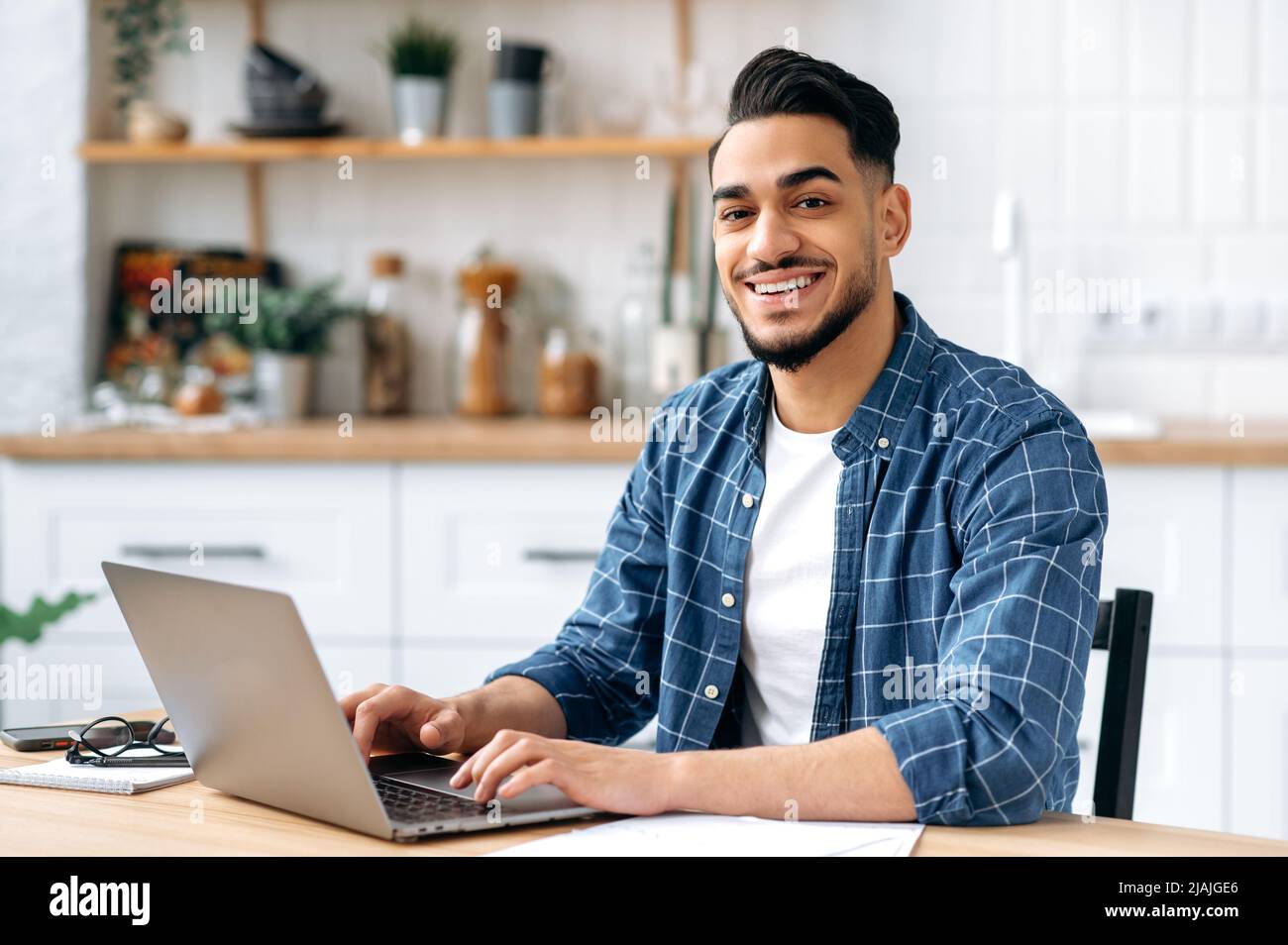 Porträt eines attraktiven positiven indischen oder arabischen Mannes, Unternehmers oder Freiberuflers, der aus der Ferne arbeitet, während er zu Hause in der Küche sitzt, einen Laptop benutzt, ein kreatives, erfolgreiches Projekt entwickelt und lächelt Stockfoto