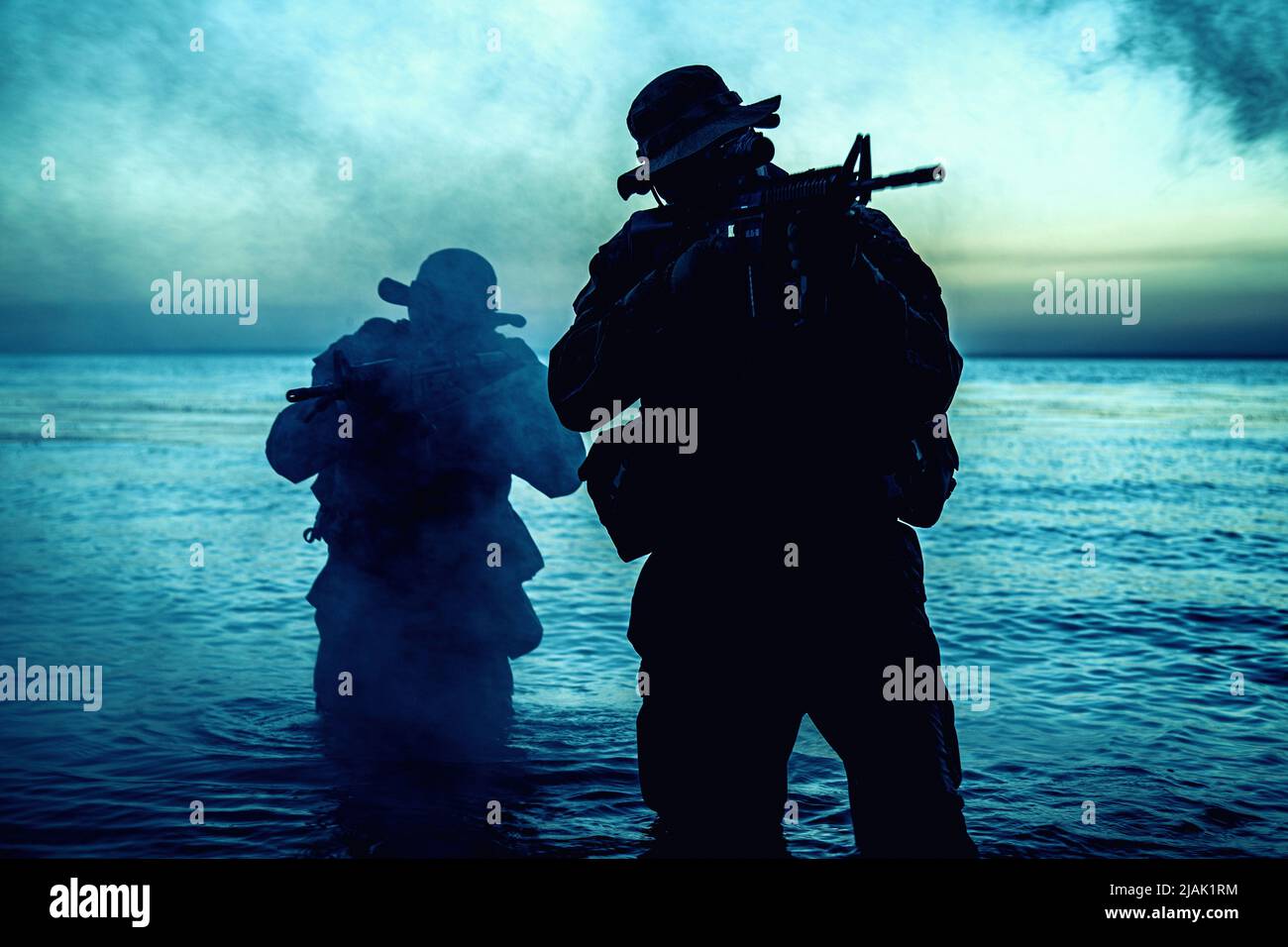 Silhouette von zwei Soldaten, die an der Küste watten und sich mit gezogenen Sturmgewehren in der Dunkelheit schleichen. Stockfoto