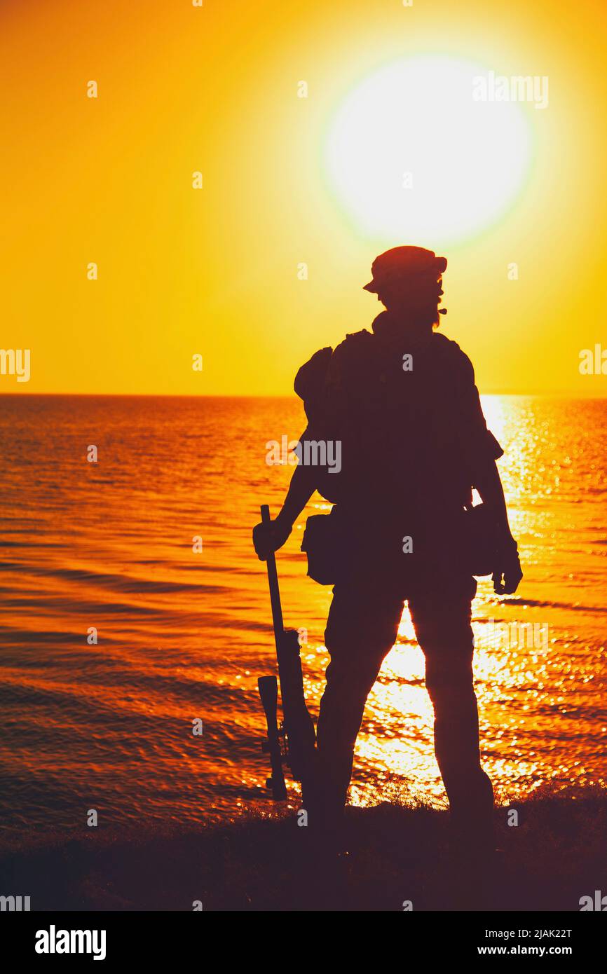 Silhouette eines Soldaten, der mit Gewehr in der Hand steht, an der Küste gegen einen Sonnenuntergang. Stockfoto