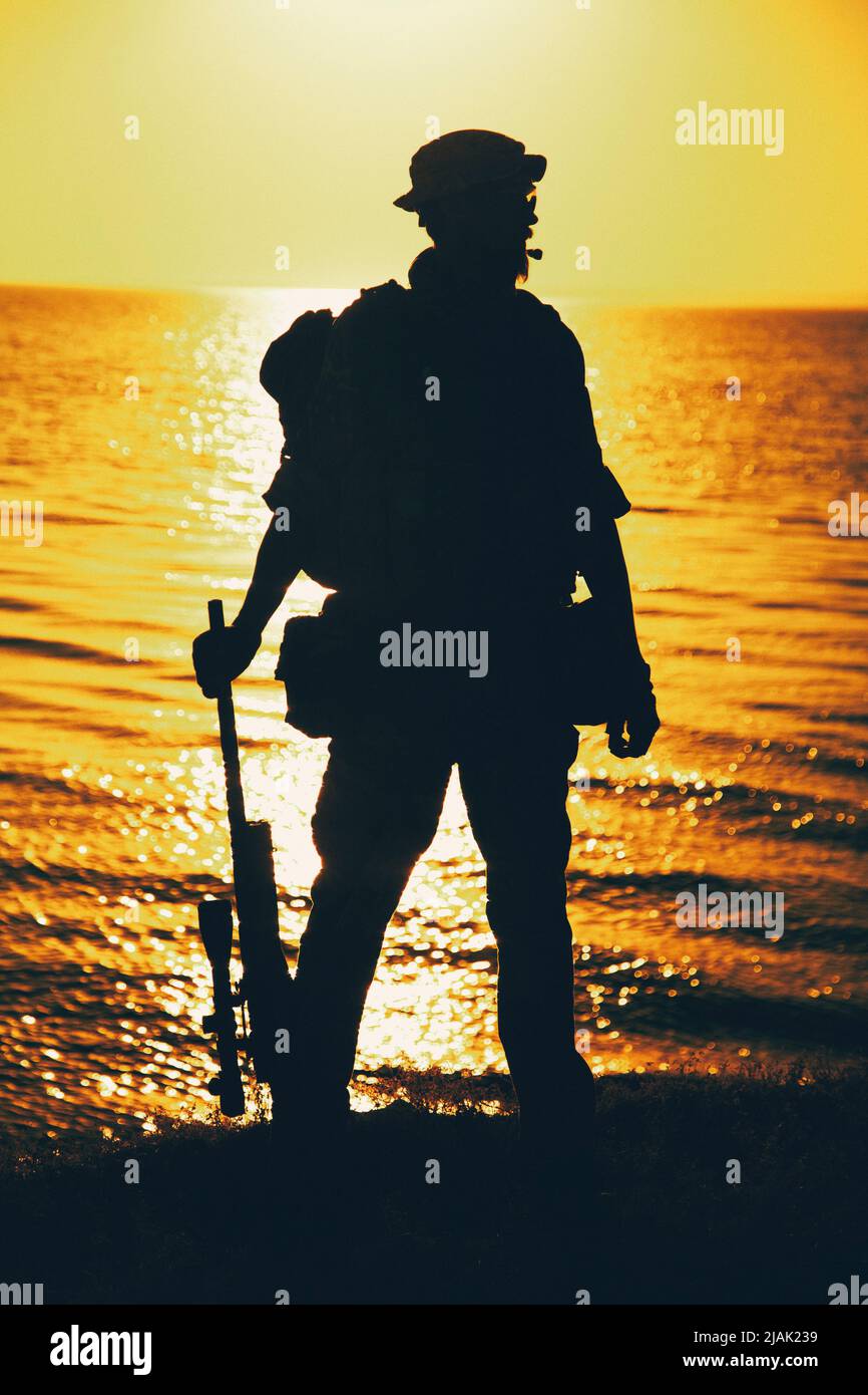 Silhouette eines Soldaten, der mit Gewehr in der Hand steht, an der Küste gegen einen Sonnenuntergang. Stockfoto