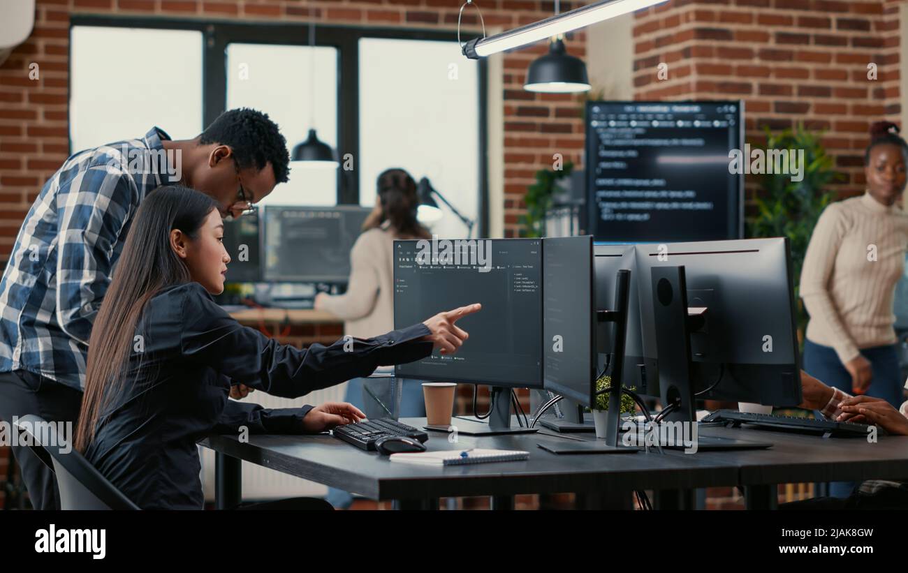 Gemischtes Team von Softwareentwicklern, die den Quellcode analysieren und auf Bildschirme zeigen, wobei der Algorithmus mit der Benutzeroberfläche auf einem digitalen Tablet verglichen wird. Programmierer, die am Codierungsgruppenprojekt arbeiten, sitzen am Schreibtisch. Stockfoto