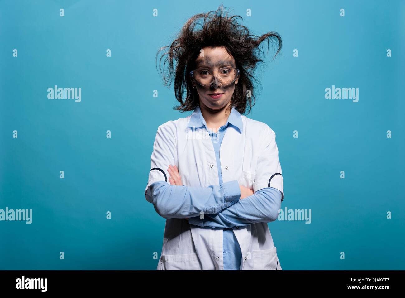 Komisch aussehende verrückte Chemikerin mit unordentlicher Frisur und schmutzigem Gesicht, das auf blauem Hintergrund steht und die Kamera anschaut. Humorvoller verrückter Wissenschaftler mit unberechenbaren Haaren und schmutzigem Gesicht, das mit gekreuzten Armen steht. Stockfoto