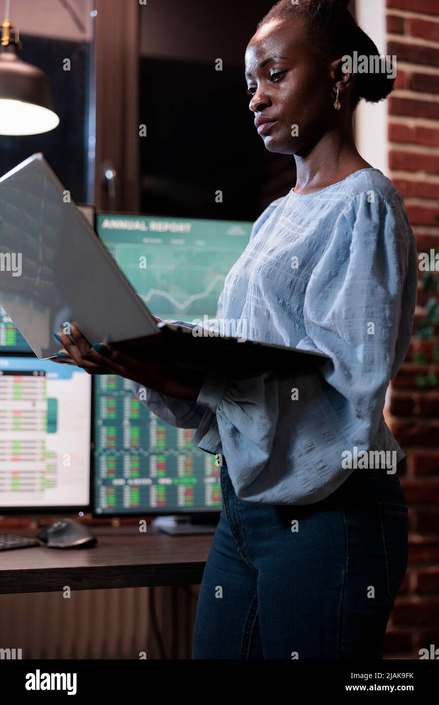 afroamerikanischer Devisenmarkthandelsagent mit einem Laptop, der vor einer Multi-Monitor-Workstation steht. Maklerfirma Mitarbeiter mit Computer während der Analyse von Live-Grafiken Stockfoto