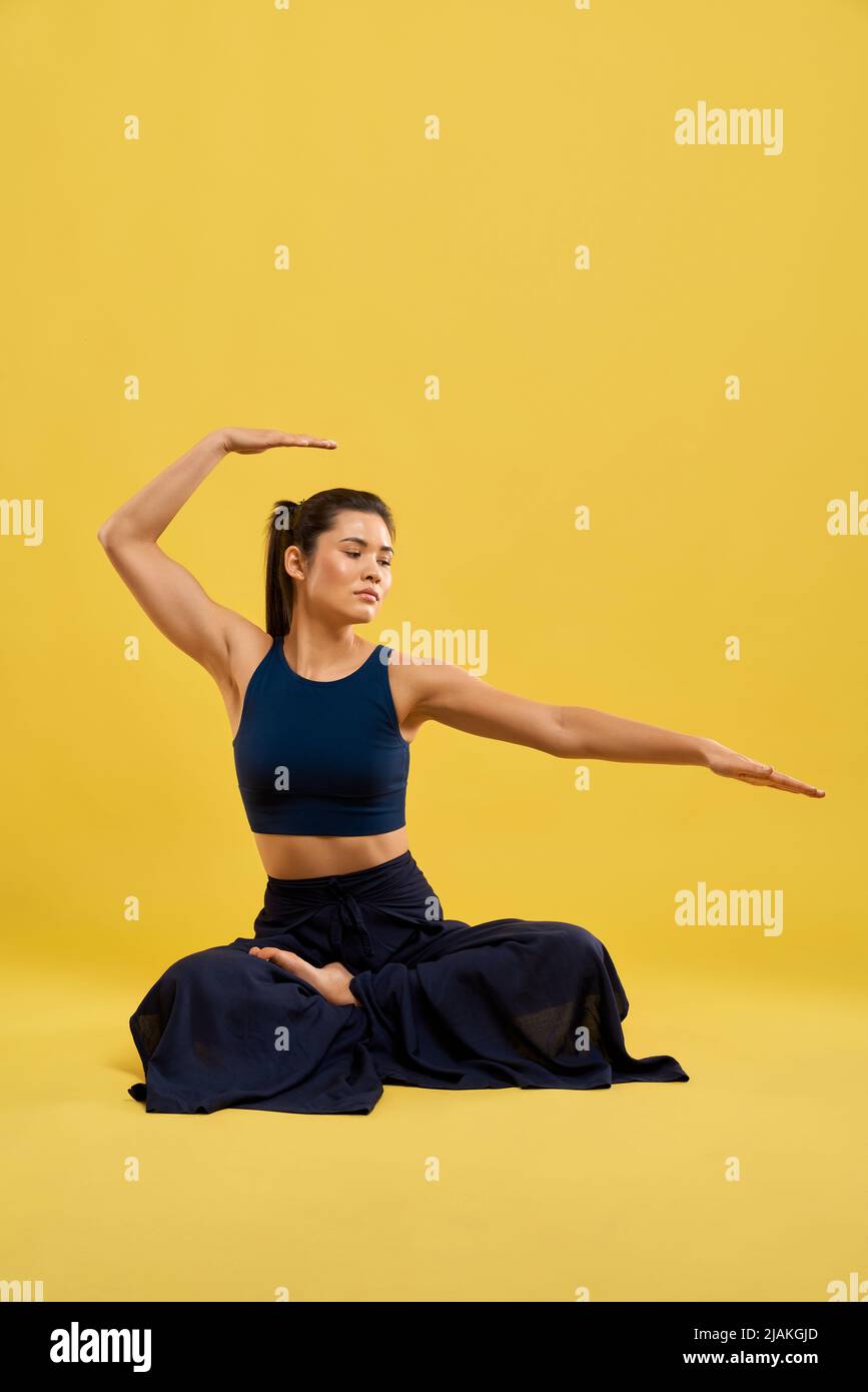 Konzentrierte Frau, die Yoga-Asana im Studio durchführt. Vorderansicht einer sportlichen Frau in Lotushaltung mit erhobener, gebeugener Hand über den Kopf und ausgestreckten Fingern während des Trainings. Konzept des Yoga Asana. Stockfoto
