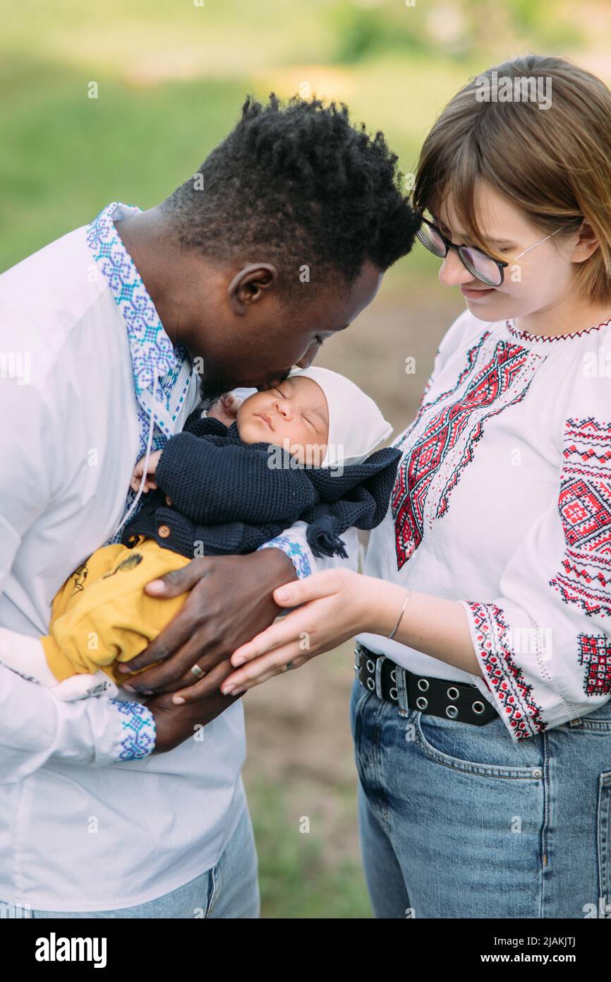 Happy interracial Familie in ukrainischen nationalen bestickten Shirts gekleidet hält und küsst ihr neugeborenes Baby. Konzept der interracial Familie und Einheit Stockfoto