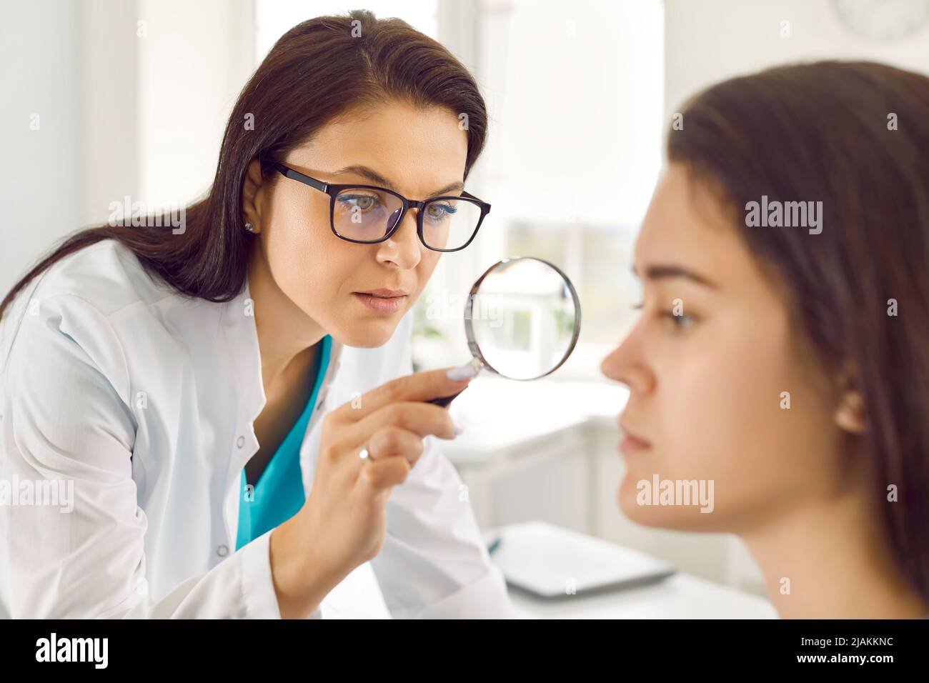 Der Dermatologe verwendet Lupe, um die problematische Haut im Gesicht einer jungen Frau zu untersuchen Stockfoto