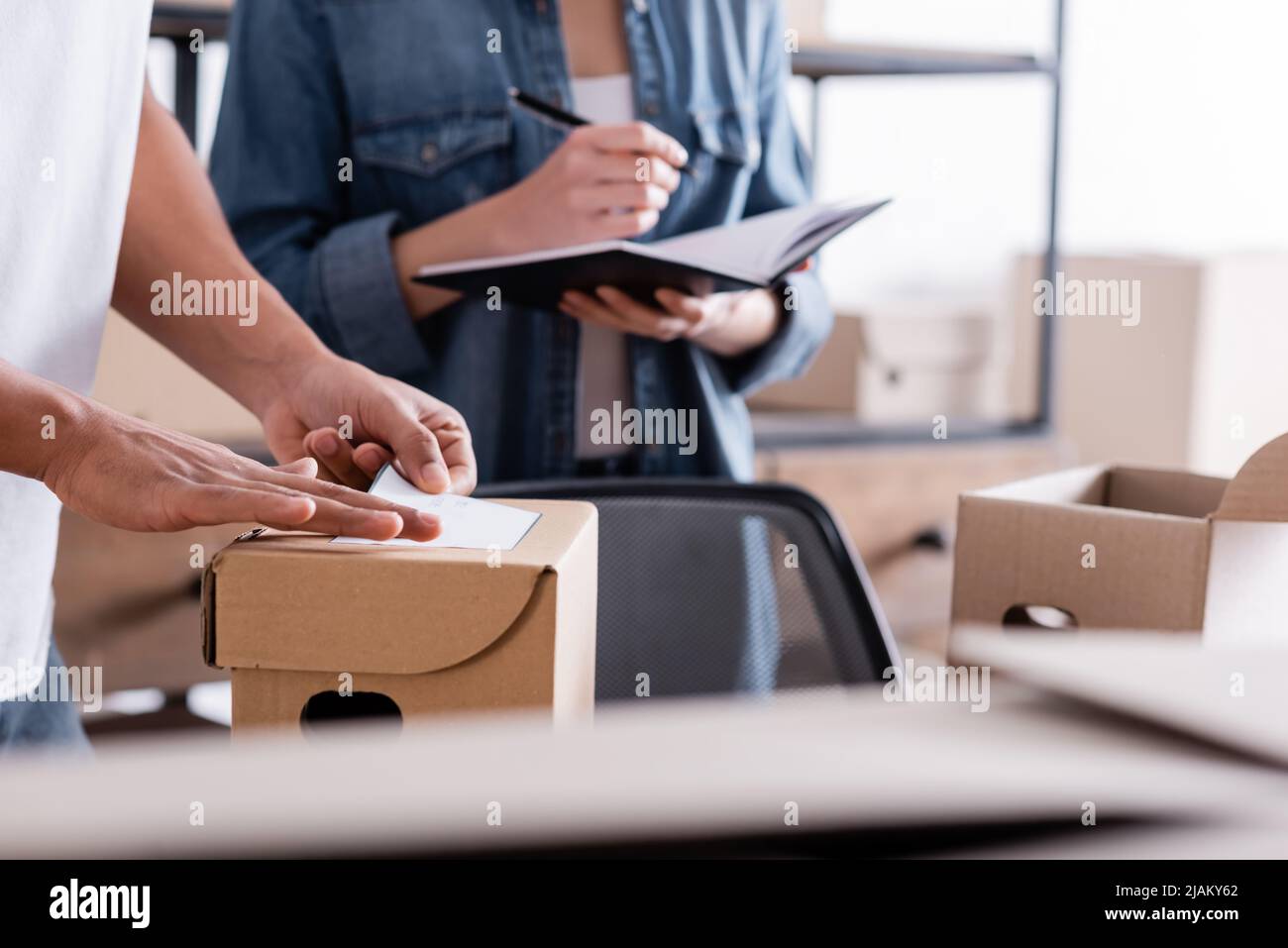 Beschnittene Ansicht des afroamerikanischen Verkäufers, der Versandetikett auf dem Paket in der Nähe eines Kollegen mit Notebook im Online-Shop angebracht hat Stockfoto