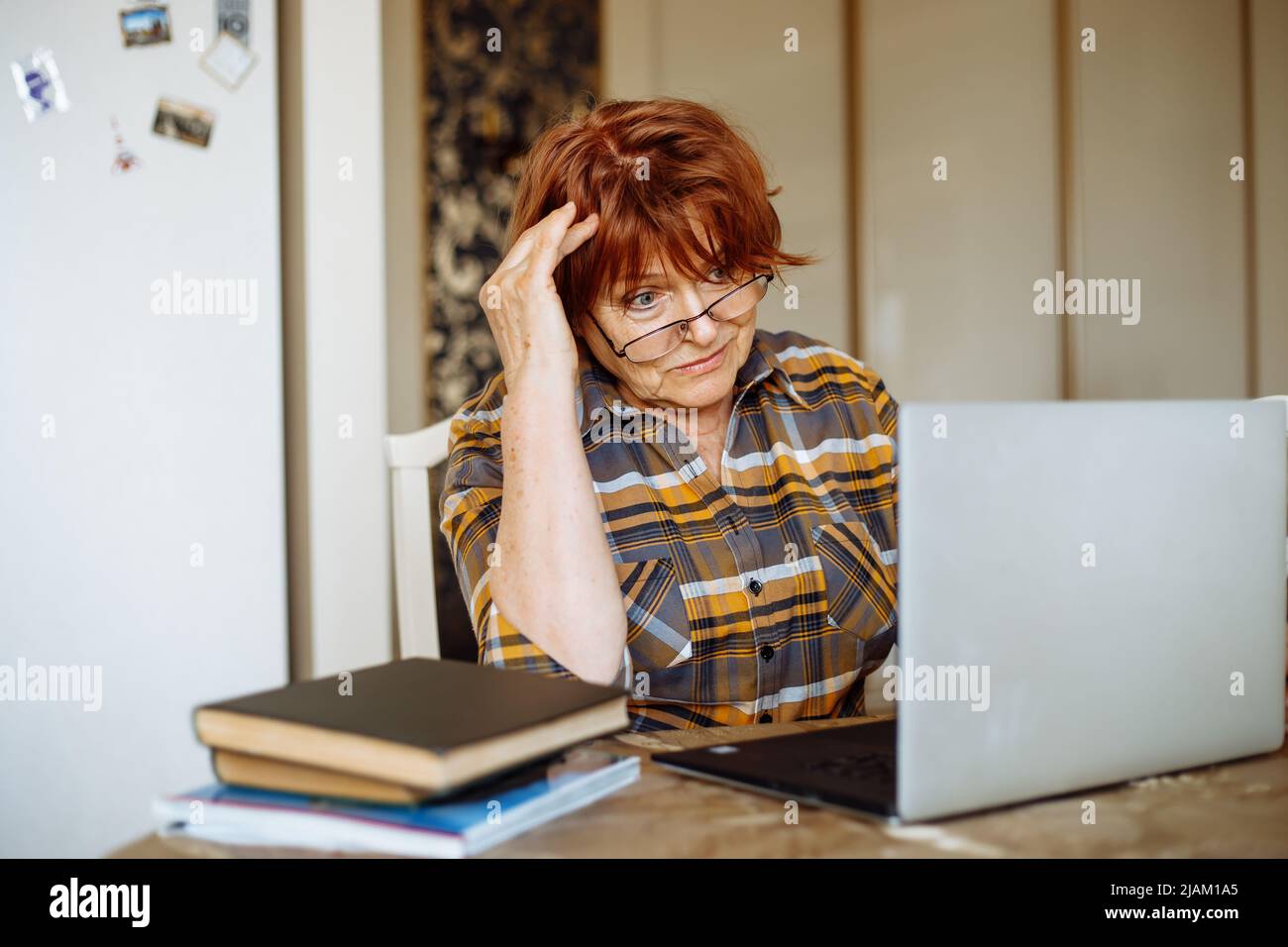 Kämpfende aktive ältere Frau in Brille mit Laptop, Online-Studie in der heimischen Küche. E-Learning, Videoanrufe Stockfoto