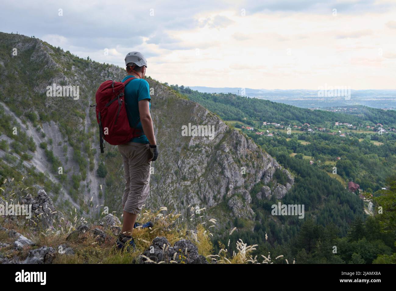 Mann mit alpinem Kletterhelm, rotem Rucksack und Dreiviertel-Sporthose, am Rande einer Bergklippe, blickt in die Ferne. Bergsteigen Co Stockfoto