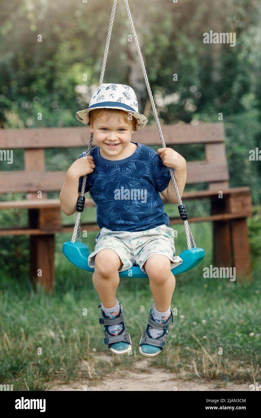 Glücklicher kleiner Junge mit Hut, der Spaß auf Schaukel im heimischen Garten hat. Gesundes Kleinkind schwingt am sonnigen Sommertag. Aktivitäten für Kinder im Freien, aktiv Stockfoto