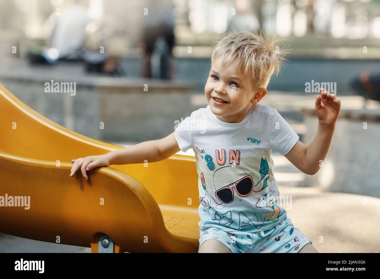 Ein fröhlicher, lächelnder, sympathischer kleiner Junge auf dem Spielplatz auf einem gelben Schieber. Das Haar des Kindes ist elektrisiert und borstend. Stockfoto