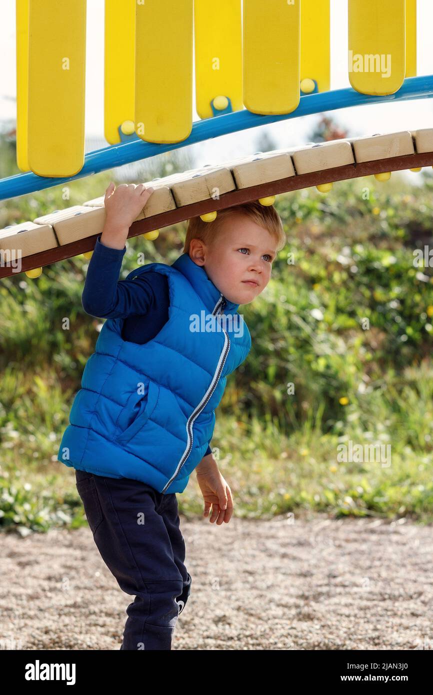 Ein kleiner niedlicher Junge spielt unter einer schönen, geschwungenen Brücke auf einem bunten Spielplatz. Ein agiles Kind verbringt aktiv Zeit an der frischen Luft. Vertikaler Phot Stockfoto