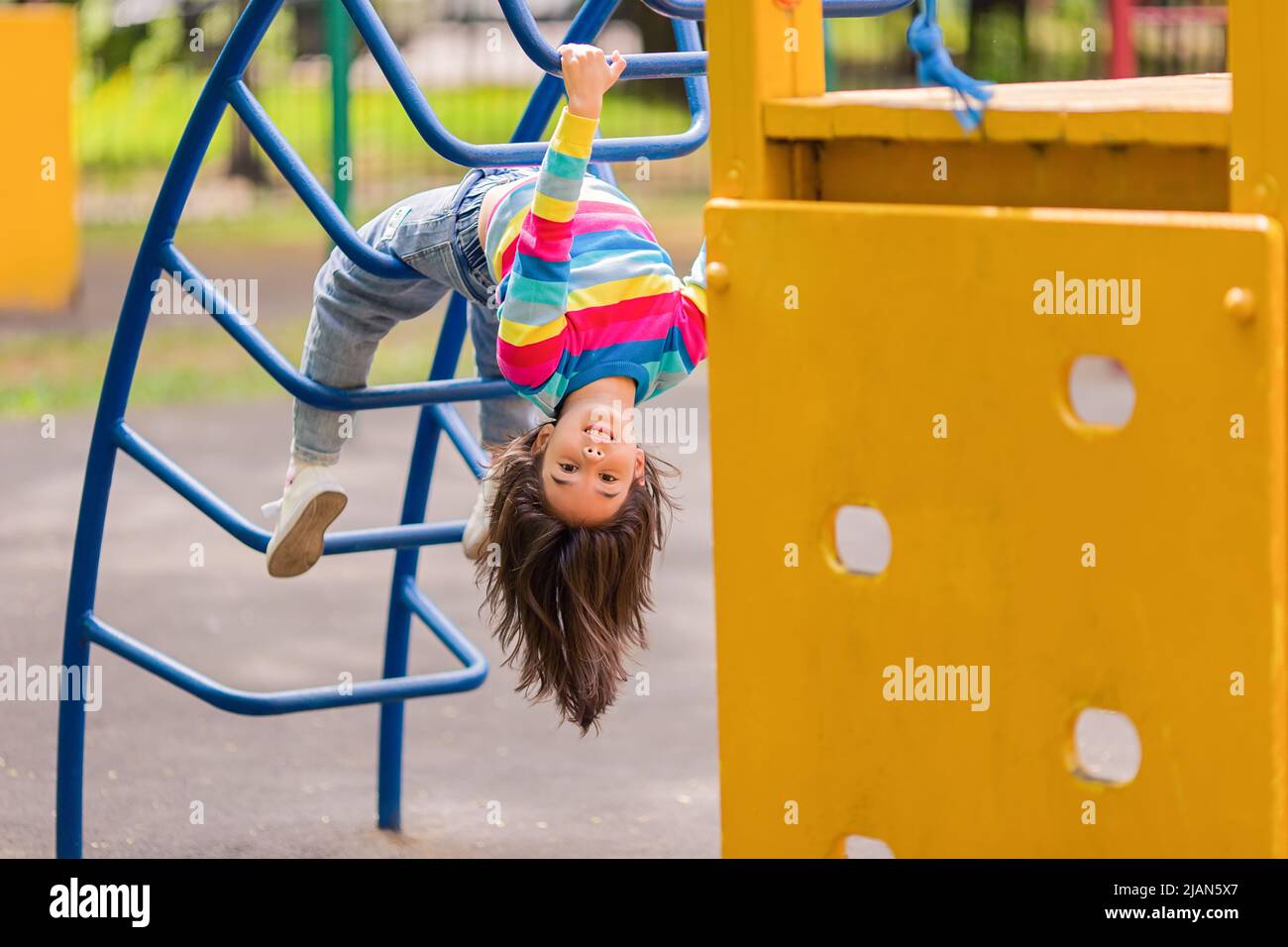 Lustige kleine lächelnde Mädchen 5-6 Jahre alt, hängend kopfüber auf der Kindertreppe auf dem Spielplatz Stockfoto