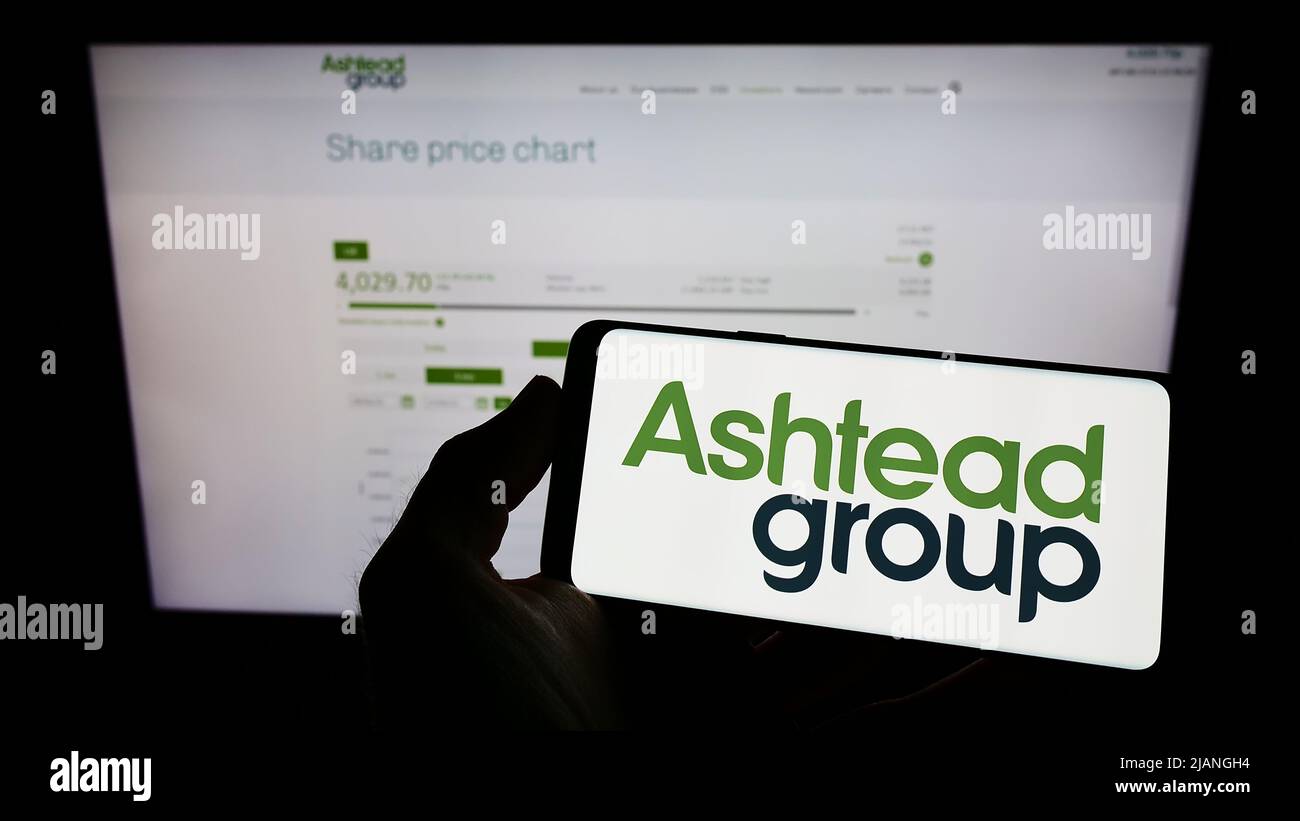 Person, die ein Mobiltelefon mit dem Logo des Ausrüstungsverleihs Ashtead Group plc auf dem Bildschirm vor der Webseite hält. Konzentrieren Sie sich auf die Telefonanzeige. Stockfoto