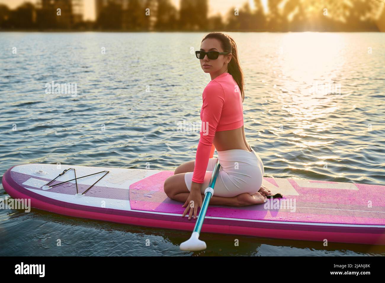 Attraktive kaukasische Frau trägt trendige Sonnenbrillen und bunte Sportkleidung üben in supboard während der Sommerzeit. Sportliche Brünette genießt aktives Hobby auf Stadtsee. Stockfoto