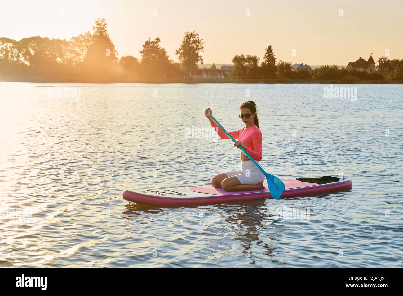 Kaukasisch fit Frau in Sonnenbrille und bunten Sport-Outfit sitzt auf supboard und rudern mit langen Paddel. Konzept der Wasseraktivität und Outdoor-Training. Stockfoto
