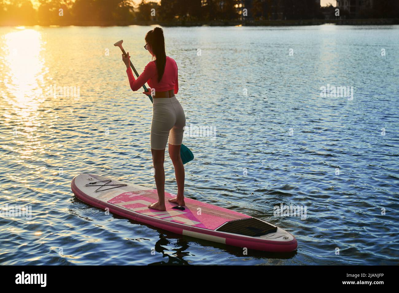 Kaukasische athletische Frau, die am Abend auf dem See im Supboarding praktiziert. Junge Brünette trägt bunte Sportkleidung. Freizeit für Wassersport im Freien. Stockfoto