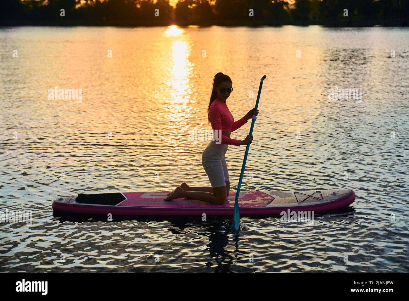 Seitenansicht der schlanken jungen Frau schweben auf supboard mit erstaunlichen Sommer Sonnenuntergang auf dem Hintergrund. Aktive kaukasische Brünette macht Abendübungen auf dem Wasser. Stockfoto