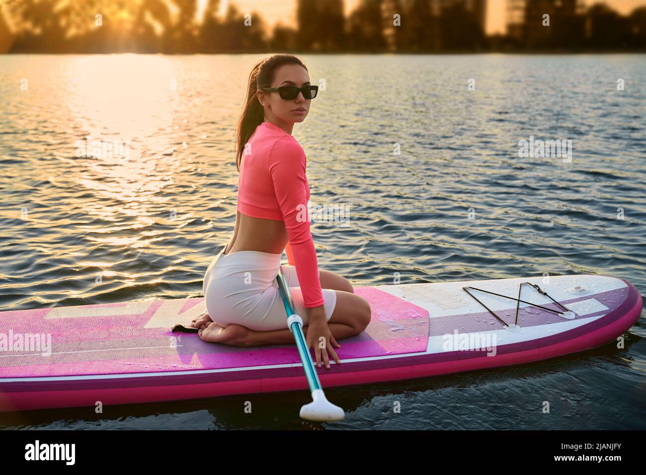 Wunderschöne junge Frau in Sonnenbrillen und Sportkleidung, die während des Sonnenuntergangs über dem See auf dem Paddle Board schwebt. Konzept eines aktiven und gesunden Lebensstils. Stockfoto