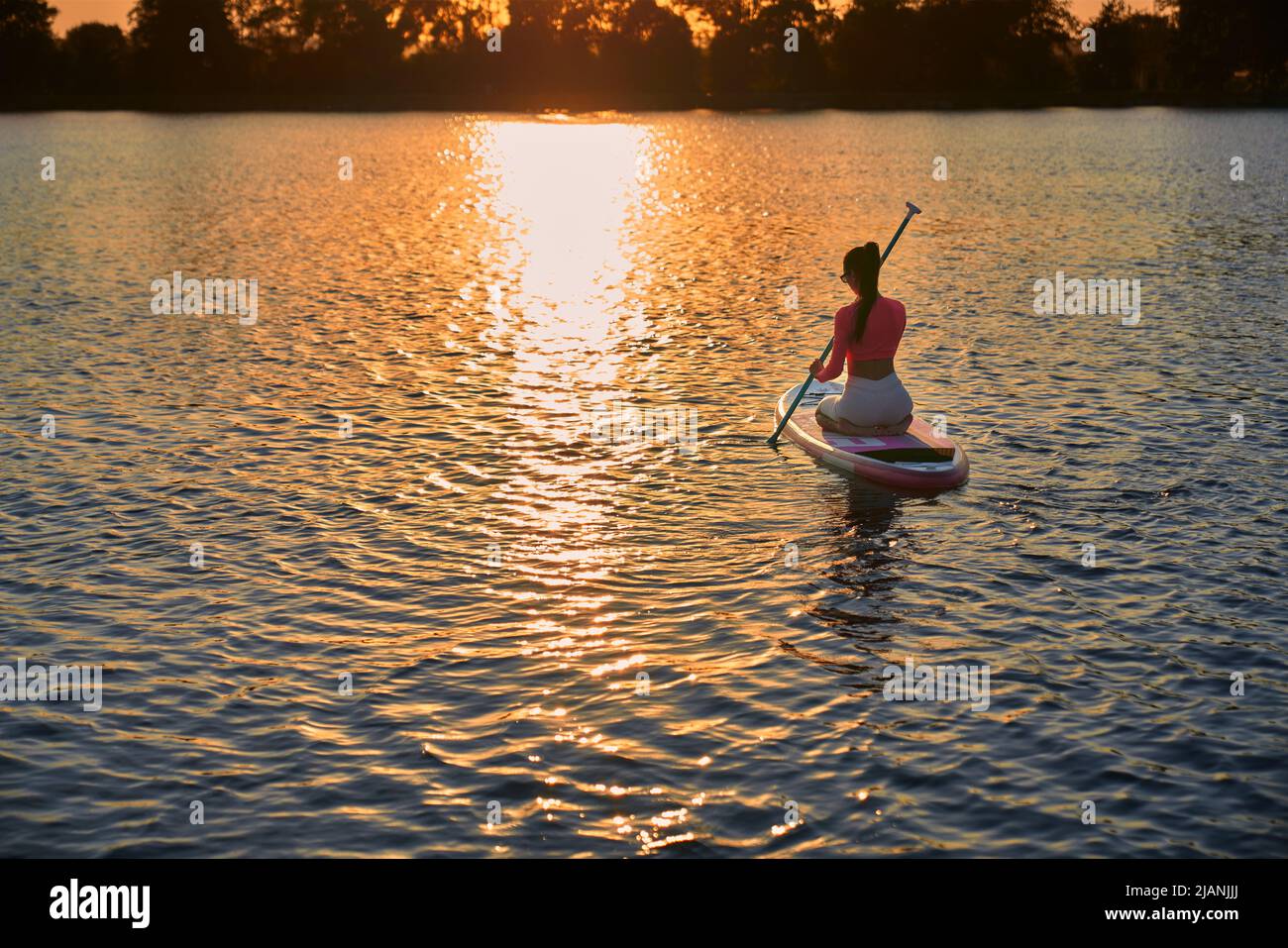 Sportliche junge Frau im aktiven Ohr, die beim SUP-Boarding den atemberaubenden Sonnenuntergang über dem See genießt. Konzept von Menschen, Freizeitaktivitäten und gesunden Lebensstile. Stockfoto