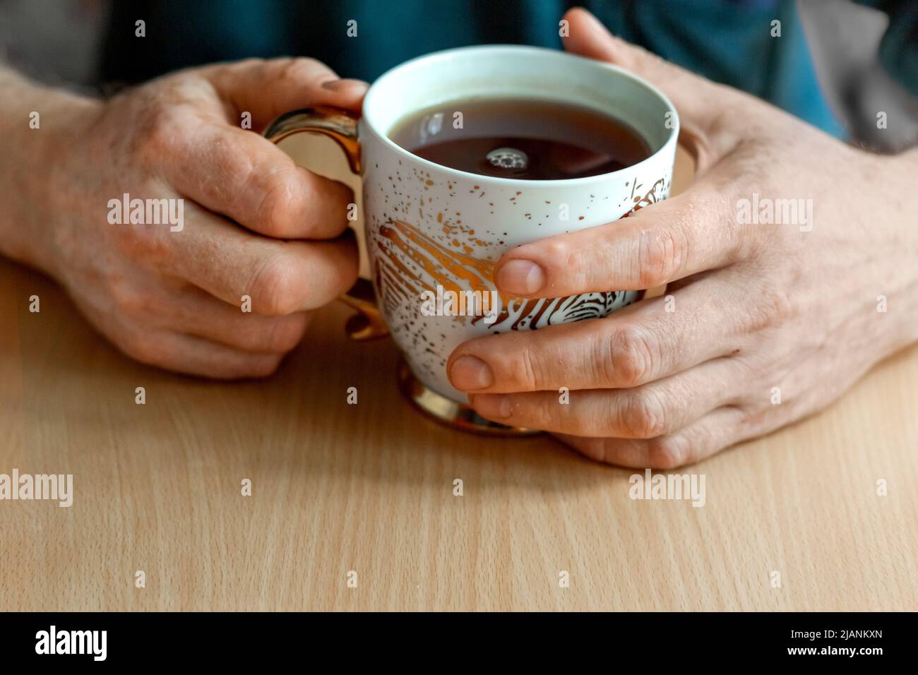 Männliche Hände mit Vitiligo-Verfärbung (fleckiger Verlust der Hautfarbe), halten eine vergoldete Tasse Hibiskus-Tee. Die Teeblätter stehen auf dem Tisch. Stockfoto