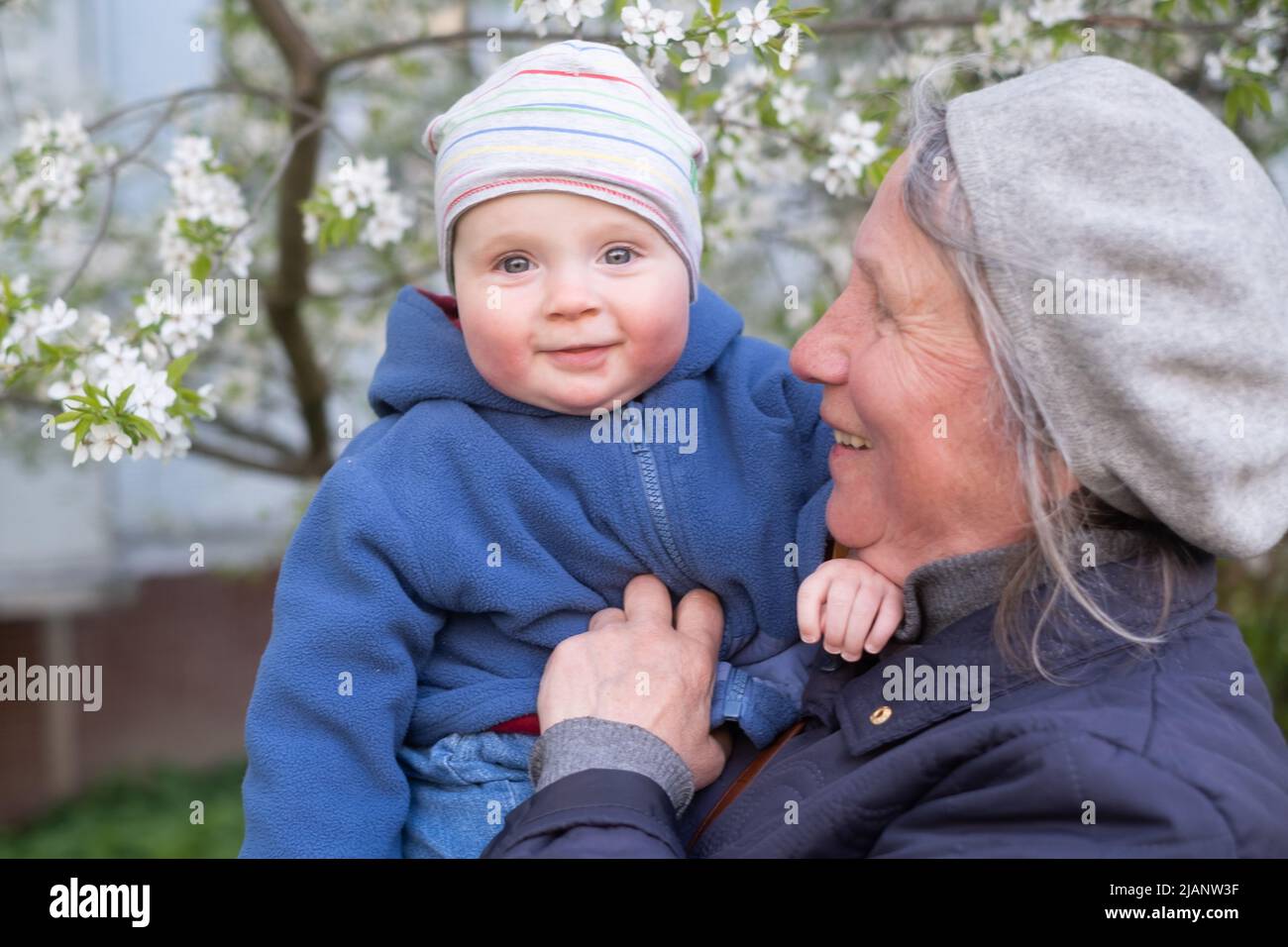 Ältere Großmutter, die ein 9 Monate altes Baby in den Armen hält und auf der Straße in der Nähe eines blühenden Baumes steht. Stockfoto