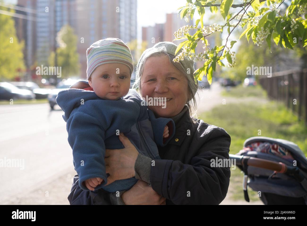 Ältere Großmutter, die ein 9 Monate altes Baby in den Armen hält und auf der Straße in der Nähe eines blühenden Baumes steht. Stockfoto