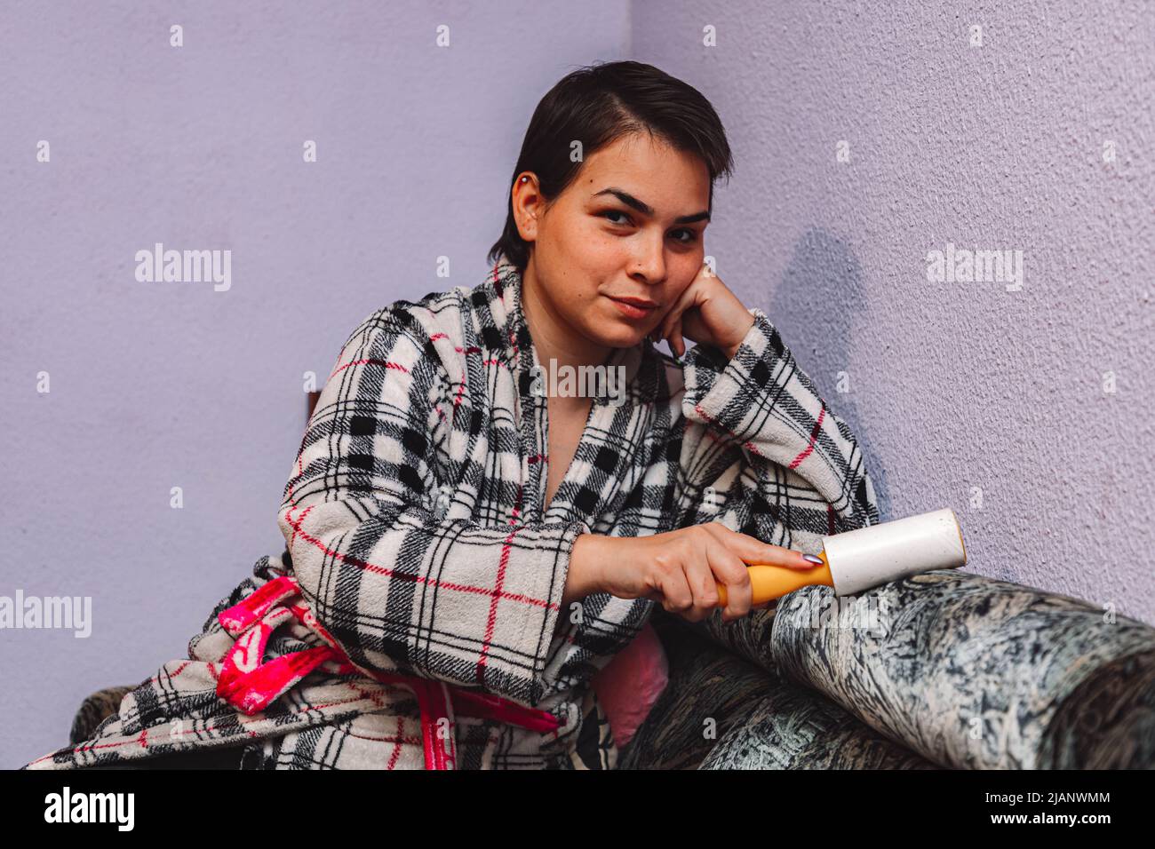 Das junge Mädchen in ihrer 20s-jährigen mit kurzen schwarzen Haaren und farbenfroher Robe reinigt das Sofa mit einem Pinsel, um Katzenhaare zu entfernen Stockfoto