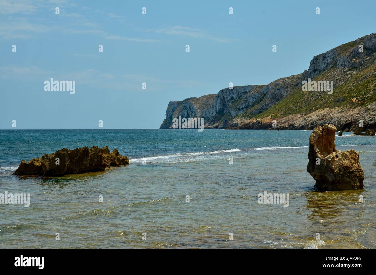 Felsen im Wasser an der Mittelmeerküste Spaniens mit Blick auf die Klippen und Berge an der Küste und das Meer unter blauem, sonnigem Himmel Stockfoto