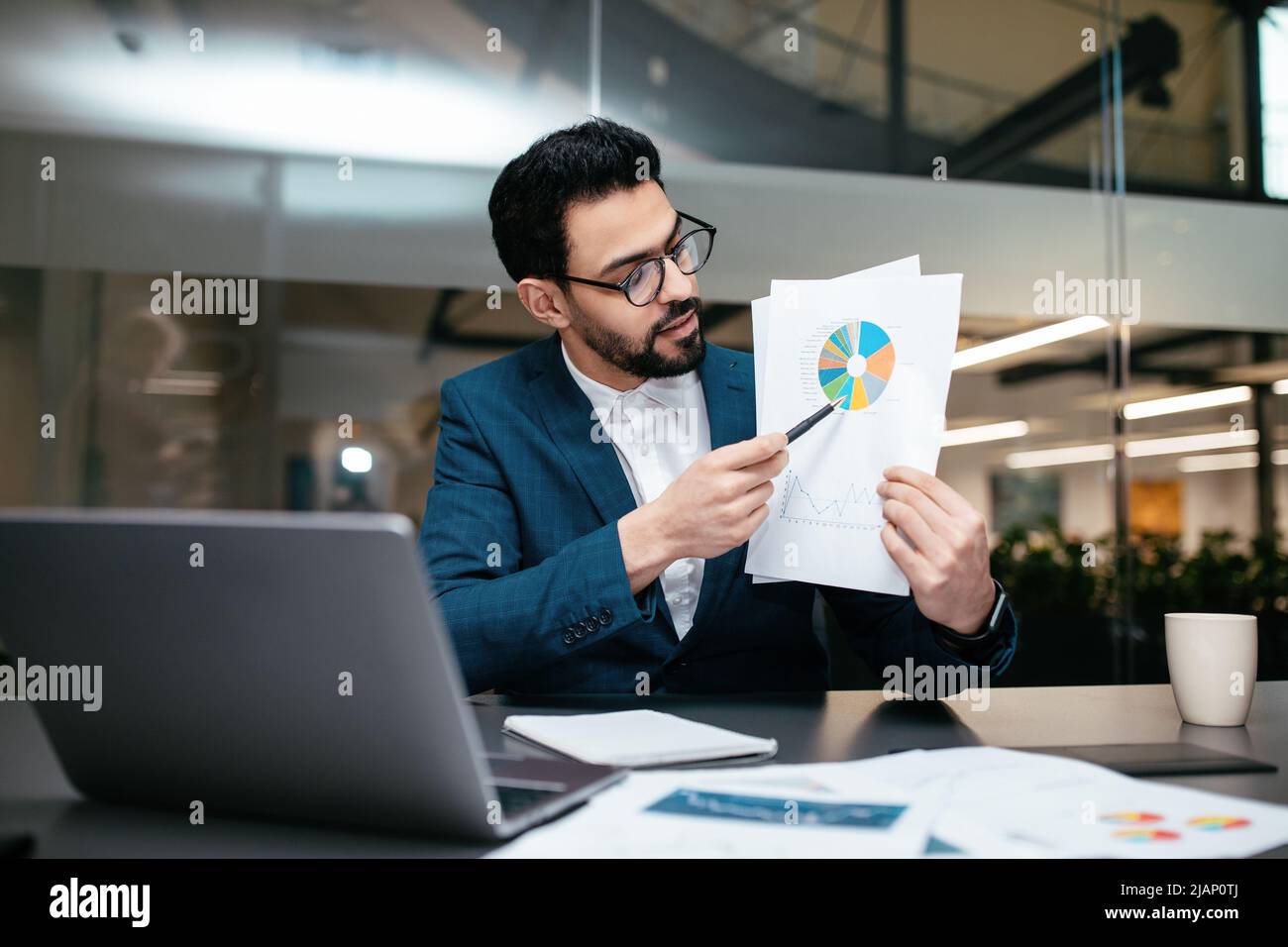 Beschäftigter junger islamischer Geschäftsmann mit Bart in Brille und Anzug zeigt Diagramm zum Laptop im Büro Stockfoto
