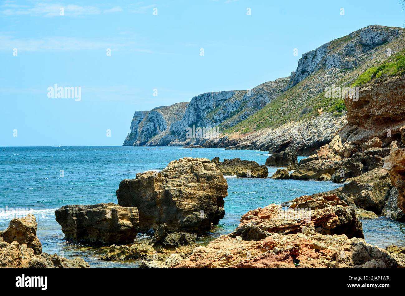 Felsen im Wasser an der Mittelmeerküste Spaniens mit Blick auf die Klippen und Berge an der Küste und das Meer unter blauem, sonnigem Himmel Stockfoto
