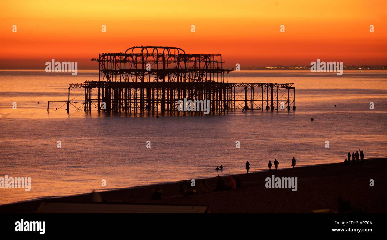 Silhouettierte Überreste des verfallenen West Pier vor einem satten orangen Sonnenuntergang. Brighton & Hove, Sussex, England, Großbritannien. Silhouetten von Menschen am Strand im Vordergrund. Stockfoto