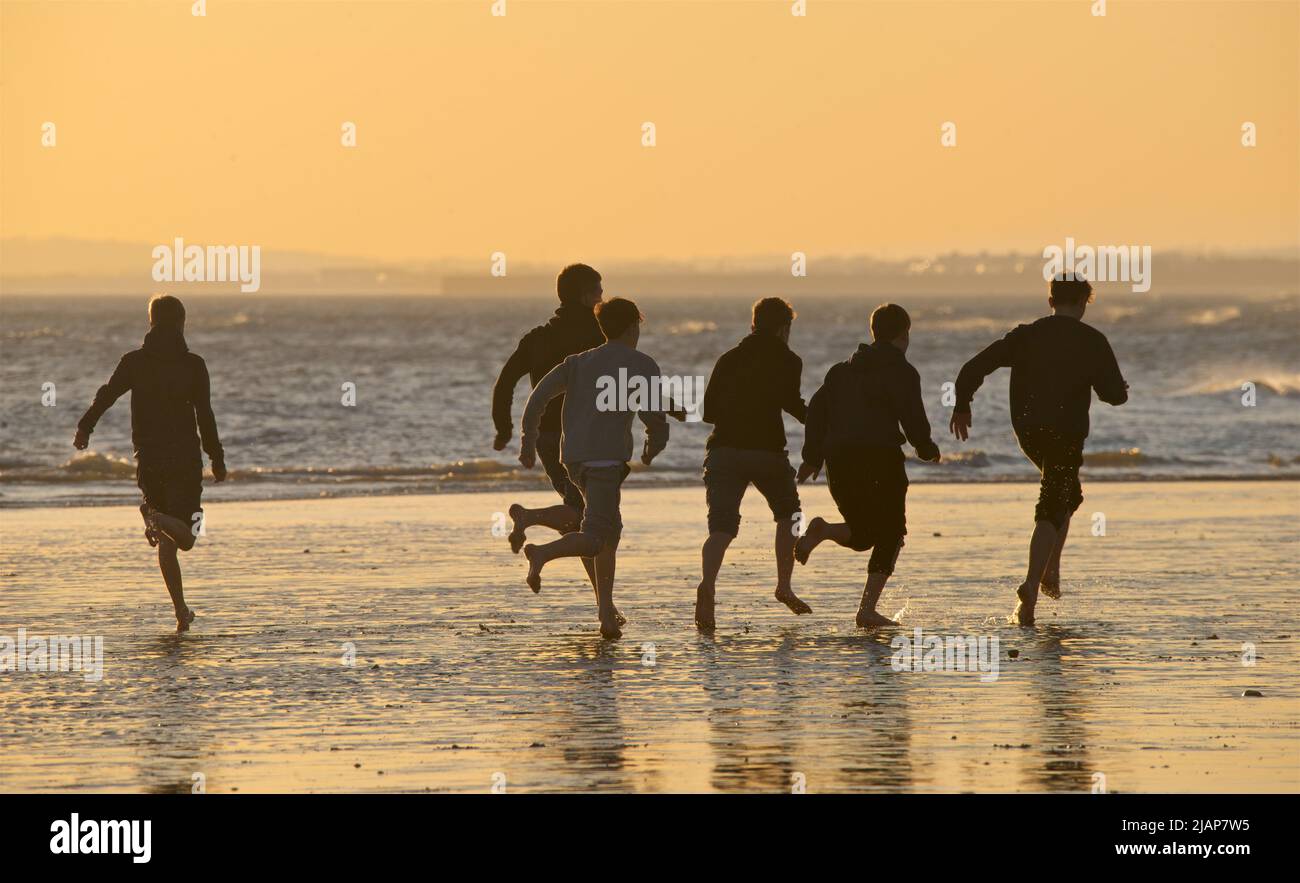 Silhouetten von Menschen am Strand bei Ebbe, Brighton & Hove, East Sussex, England, Großbritannien. Eine Gruppe von Teenagerts läuft barfuß. Stockfoto