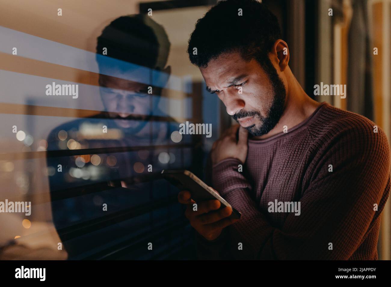 Ein junger Mann mit Smartphone sieht besorgt aus und steht abends zu Hause auf dem Balkon. Stockfoto