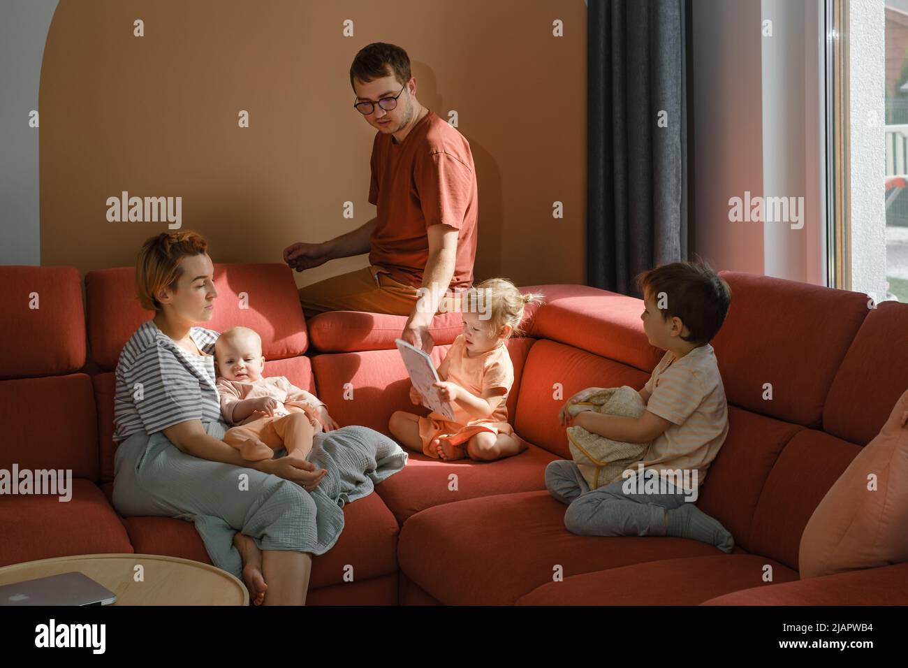 Große Familie auf der Couch zu Hause. Eltern mit Kindern auf dem Sofa während eines sonnigen Tages verbringen Freizeit. Mutter und Vater mit drei Kindern, die Buch lesen. Modern Stockfoto