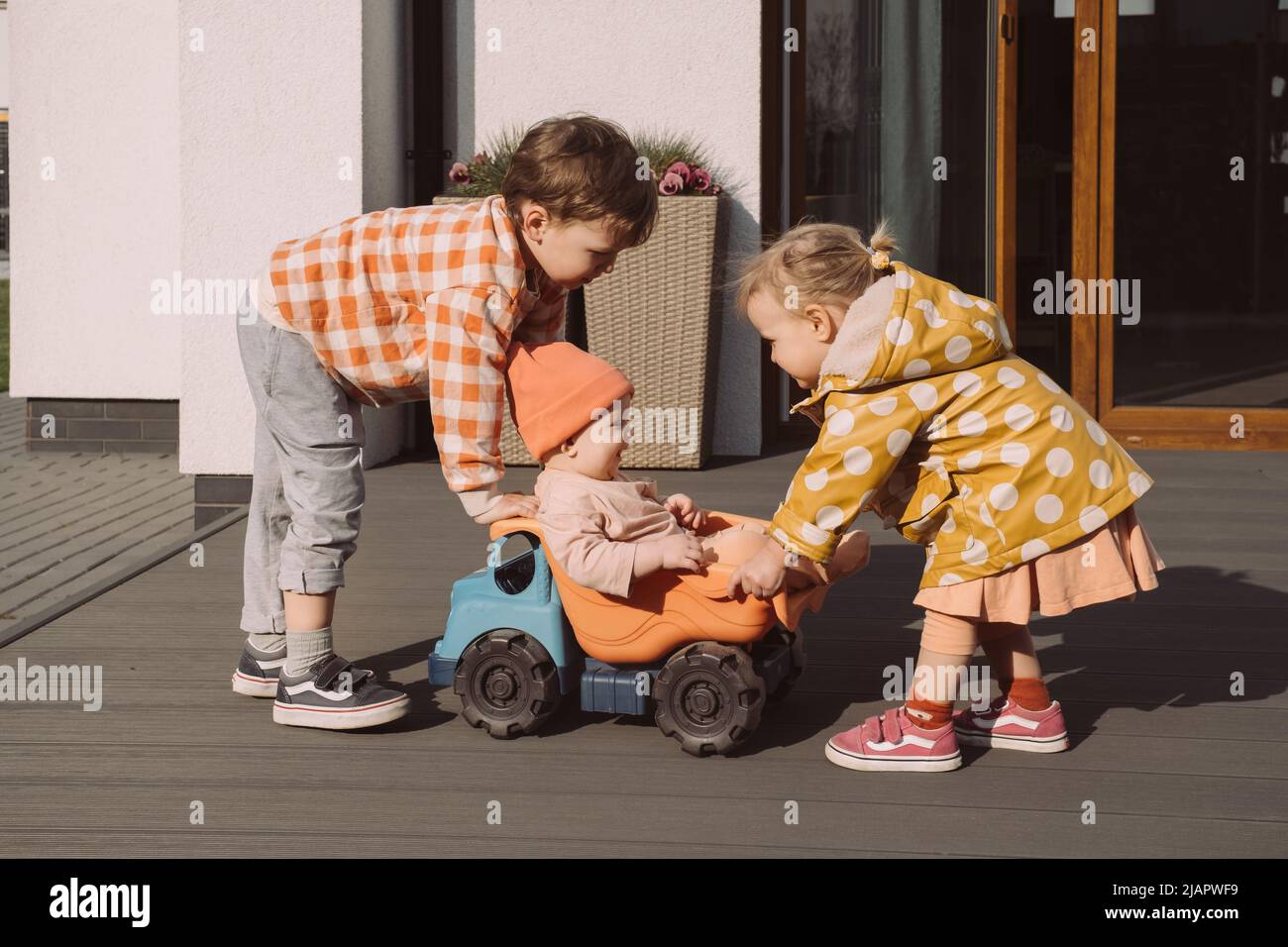 Bruder Schwester gibt eine Fahrt zu Baby Schwester in großen Spielzeugauto im Freien. Kinder haben Spaß in der Nähe des Hauses. Junge und Mädchen Rennen mit Vergnügen. Kinder Spiele von Stockfoto