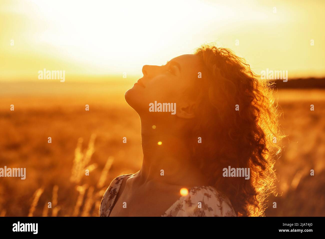 Junge marokkanerin mit braunen lockigen Haaren steht auf einem Weizenfeld, während die Sonne im Hintergrund untergeht und die Kamera blendet Stockfoto