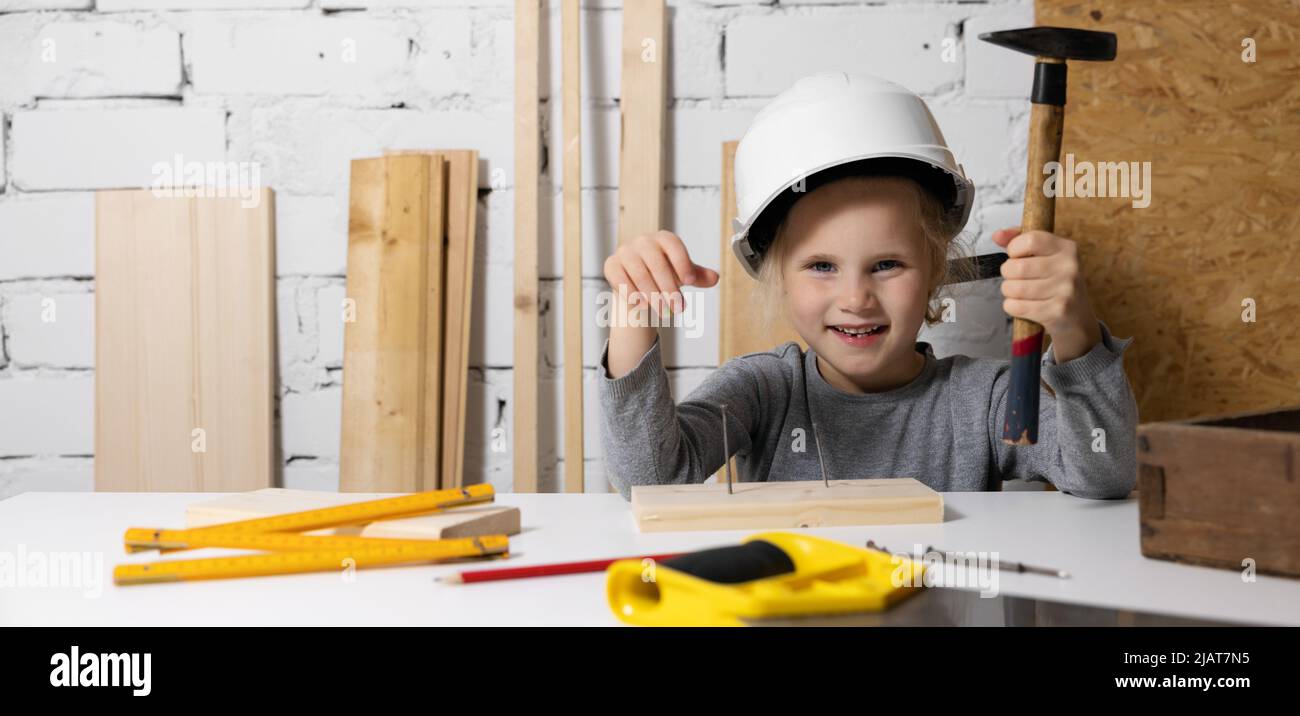 Lächelndes kleines Mädchen mit Helm lernen, einen Nagel in Holzdiele in der Holzwerkstatt zu hämmern Stockfoto