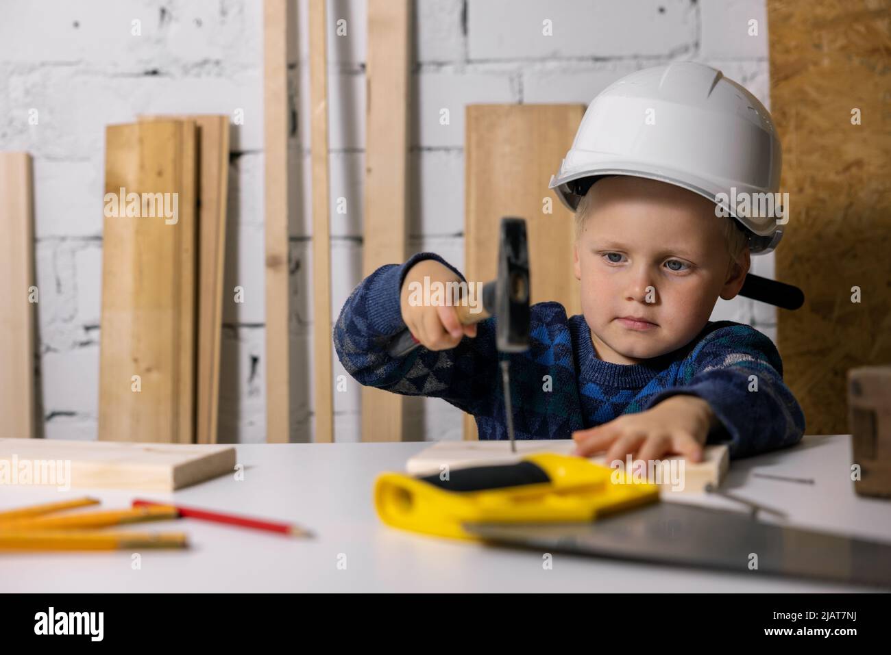Der kleine Junge mit Helm lernt in der Tischlerwerkstatt, einen Nagel in die Holzdiele zu schlagen Stockfoto
