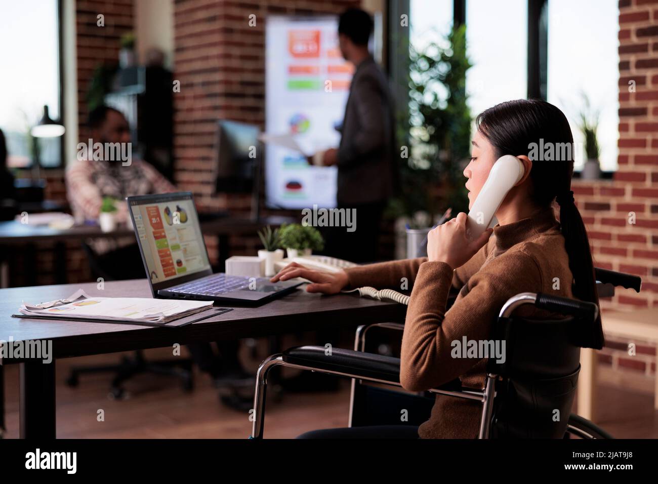 Weibliche Angestellte mit Gesundheitszustand, die Festnetzanruf in einem behindertengerechten Büro entgegennimmt. Telefongespräche über Ferngespräche, die an chronischen Beeinträchtigungen im Rollstuhl leiden. Stockfoto