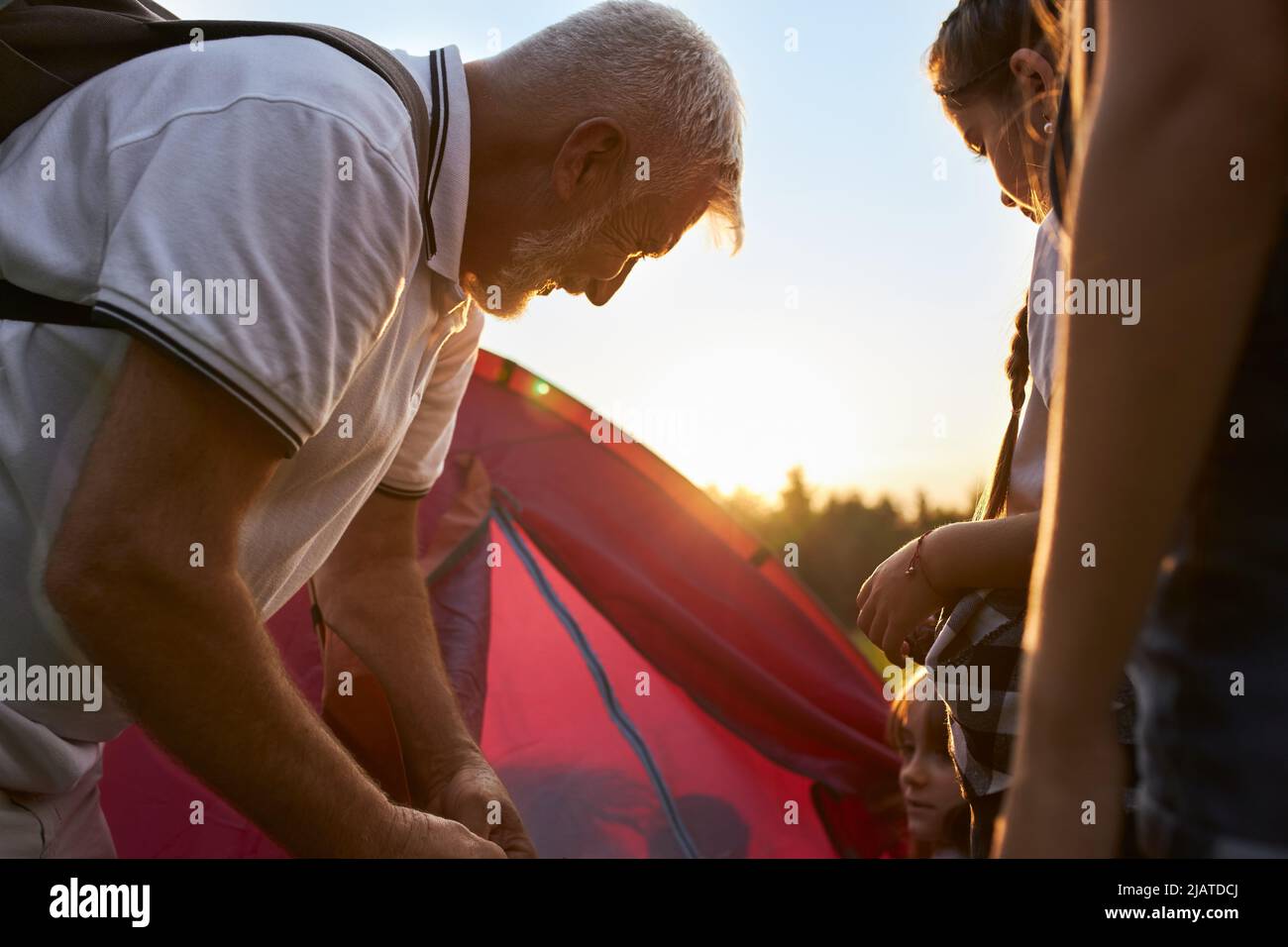 Nahaufnahme von Kindern mit einem alten Mann, der draußen ein rotes Zelt aufgeschlagen hat. Lehrer mit grauen Haaren wandern, reisen, Campingplatz zusammen mit Kindern organisieren. Konzept der aktiven Ruhe. Stockfoto