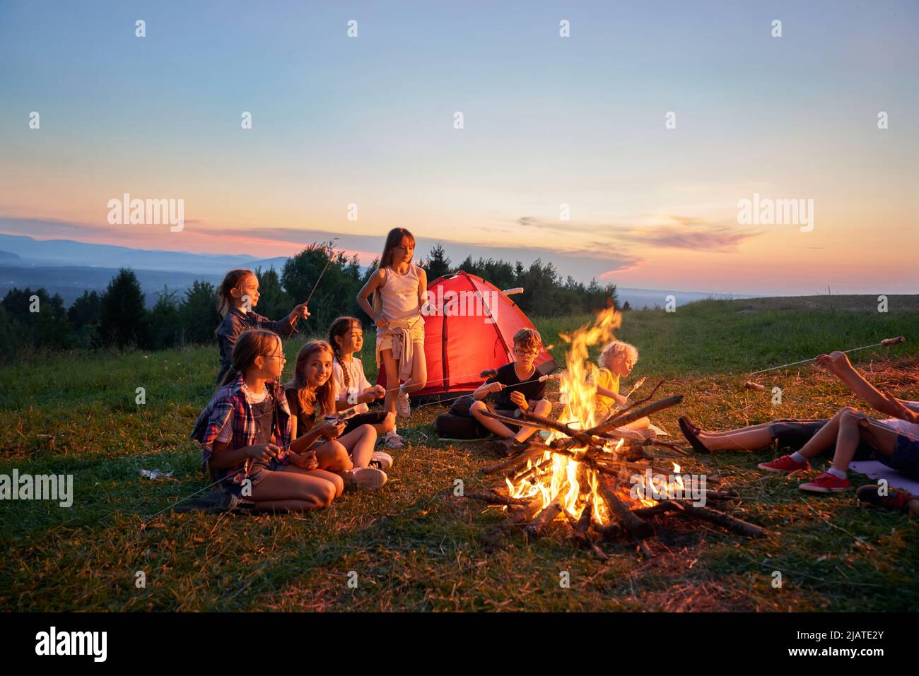 Seitenpanorama der Gruppe von Kindern, die auf dem Campingplatz in den Bergen sitzen. Kinder essen, essen, kommunizieren, sich in der Nähe des Kamins ausruhen und mieten. Konzept der aktiven Ruhe. Stockfoto